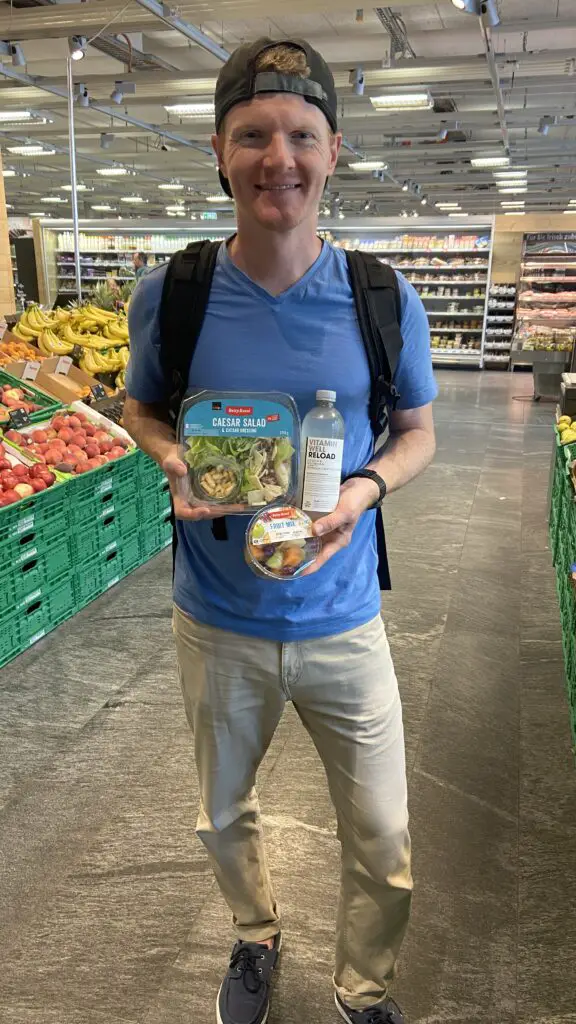 brett holding a caesar salad, fruit salad, and vitamin water from coop grocery store switzerland